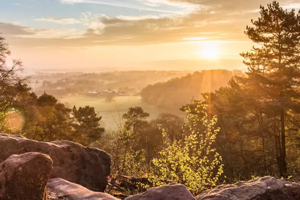 Golden sunrise over Alderley Edge in Cheshire, with soft sunlight filtering through trees and mist over the rolling countryside.