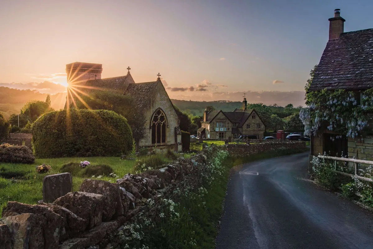 Golden sunburst shining over the historic stone church and cottages of Snowshill village in the Cotswolds.