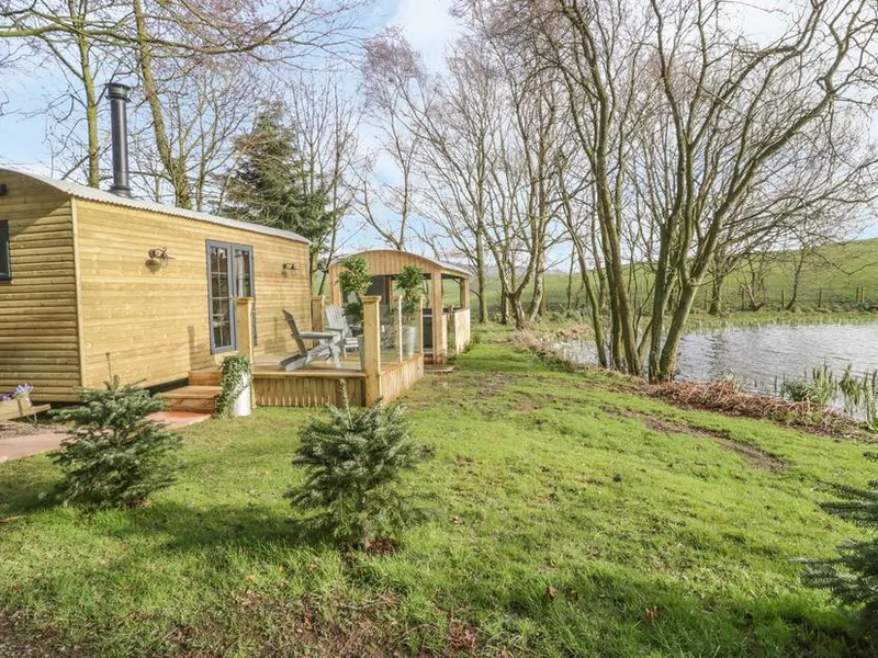 High Rigg Shepherd's Delight wooden lodge beside a lake with deck chairs and trees nearby