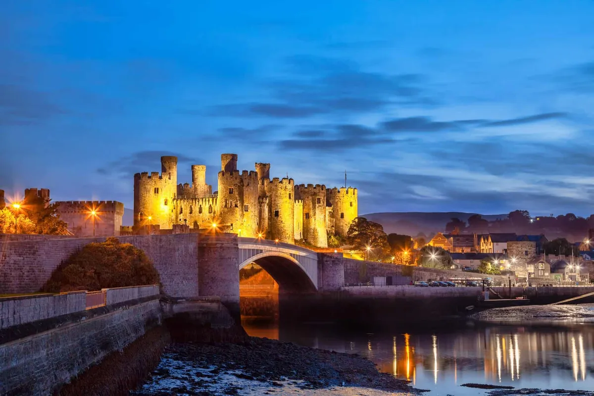 The historic Conwy Castle glows with warm lights at dusk, reflecting in the tranquil water.