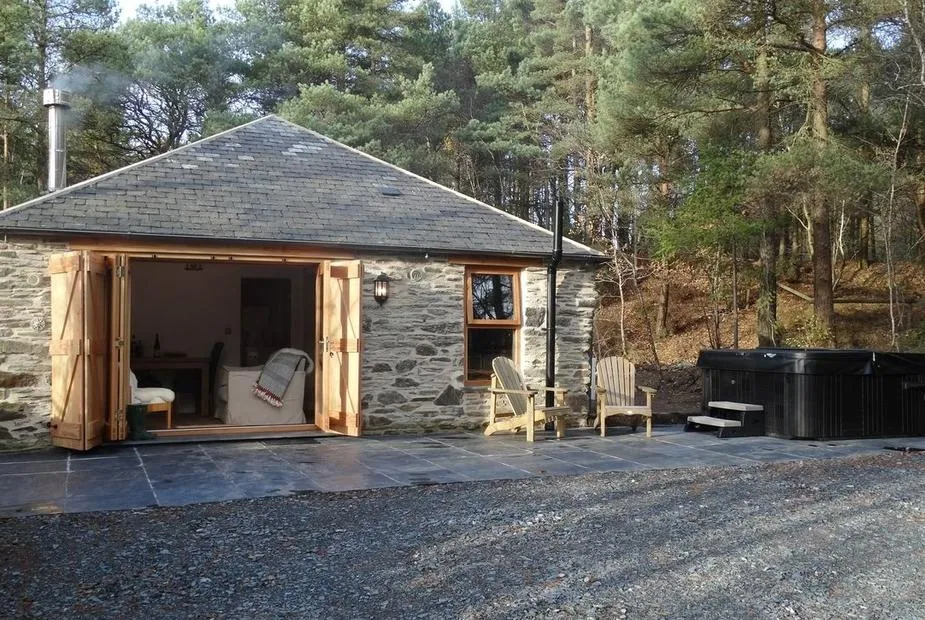 Rock View Cottage with stone exterior, open doors, patio seating and outdoor hot tub in woodland.