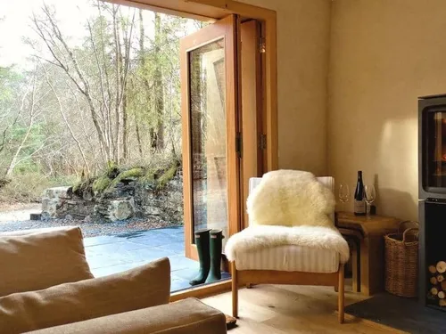 Cottage lounge with chair, wood burner, and open doors to patio and woodland