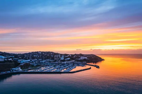 Aerial view of Torquay Marina at sunrise, with golden light reflecting off the calm sea and luxury yachts moored in the harbour.