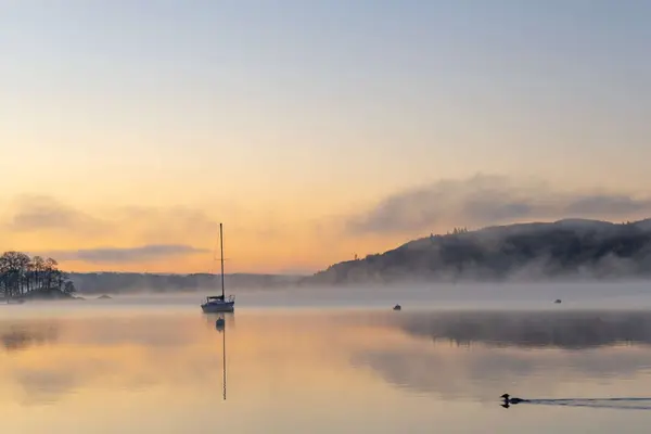 Sunrise over Lake Windermere with a lone sailboat drifting in the morning mist.